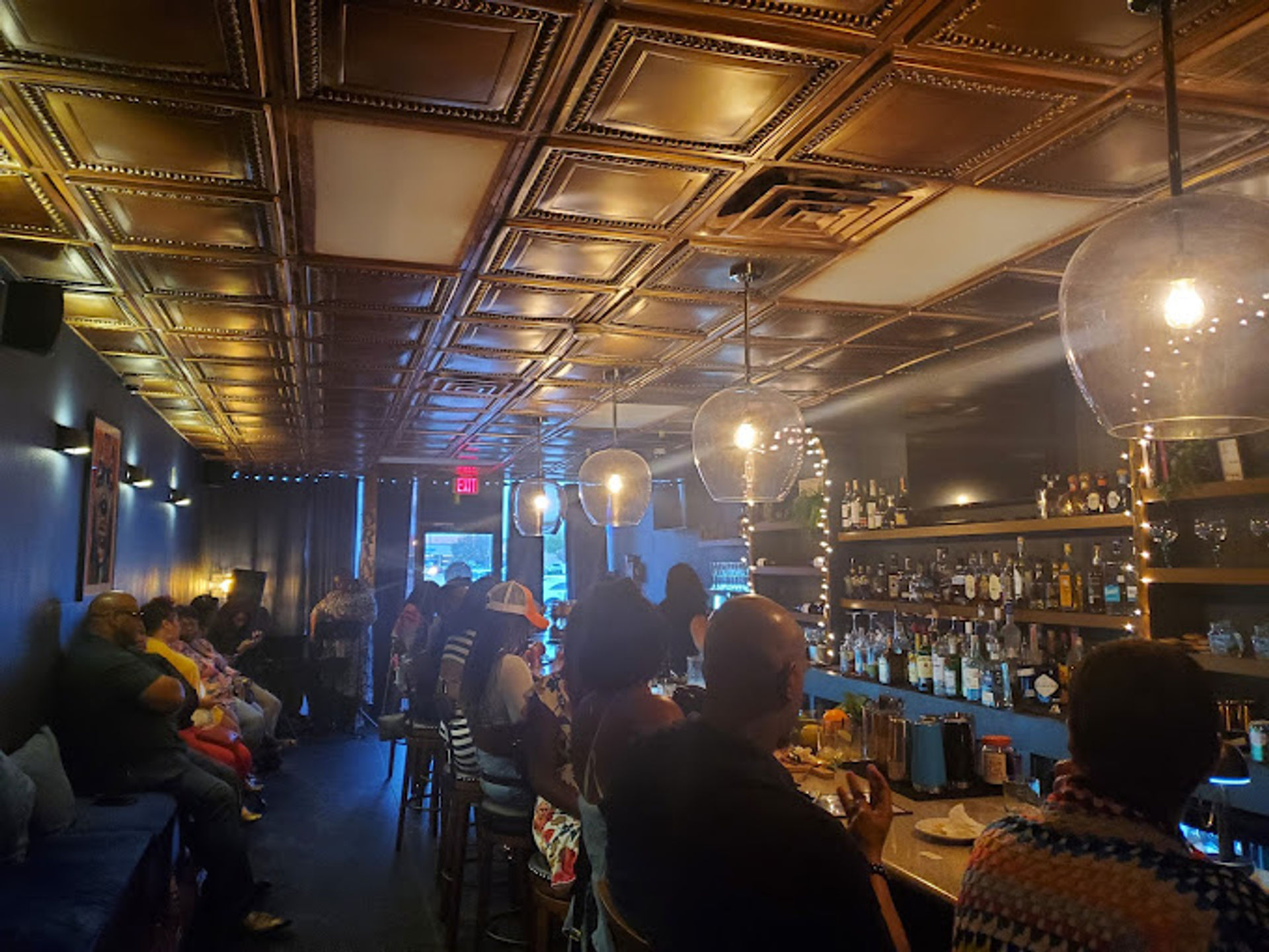 Cozy dimly lit neighborhood bar interior with vintage tin ceiling, globe pendant lights, bustling evening crowd seated at the bar and banquette, and backlit liquor shelves.