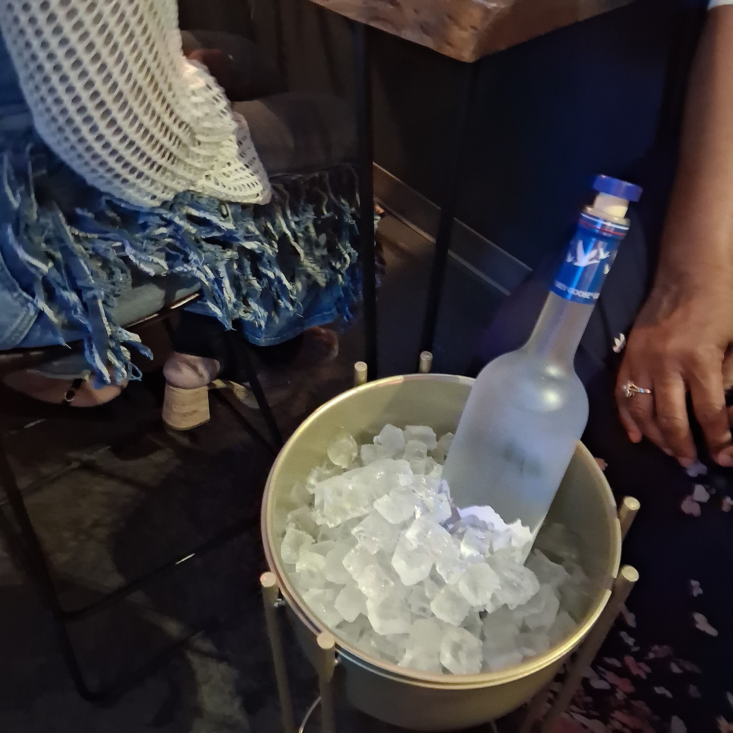 Ice bucket filled with crushed ice and a frosted spirit bottle with scoop, next to a hand with rings and a fringed denim scarf and knit shawl at a bar