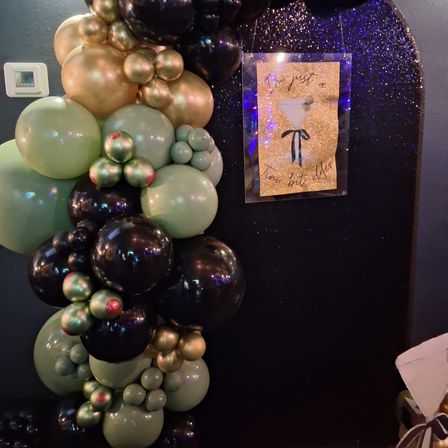 Indoor event decor: black glitter backdrop with a green, gold and black balloon garland and a sparkling martini sign — party photo booth setup.