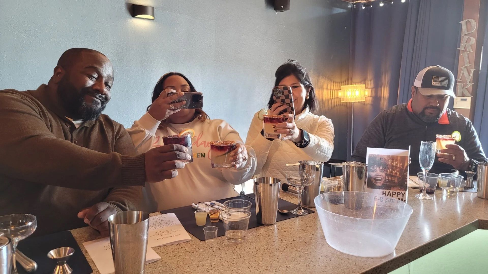 Four adults at a bar counter toasting cocktails and taking photos in a cozy indoor lounge—cheers!