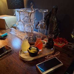 Cozy indoor bar table with upside-down wine glasses on a metal rack, three small carafes of colorful mixers, a bowl of pickles, a reserved seating sign and phones on a wooden surface.