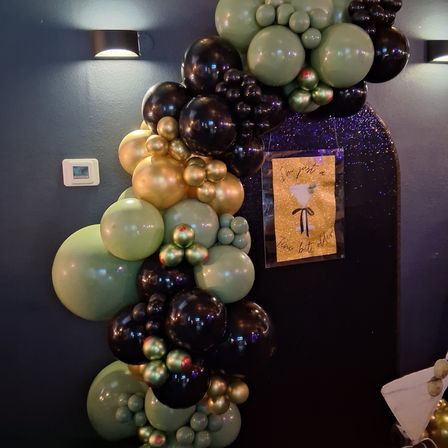 Party-ready balloon garland of black, olive green and metallic gold spheres clustered vertically against a dark gray wall with wall sconces and a glitter cocktail sign