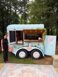 Pastel-blue mobile snack trailer serving dips under string lights in a leafy wooded park, with a smiling server beside the display.