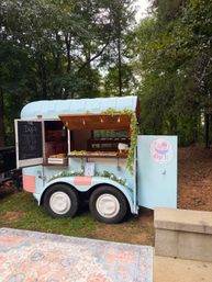 Quirky pastel-blue mobile snack trailer parked in a leafy outdoor setting, open wooden awning with string lights, chalkboard menu and trays of dips and treats on the serving counter