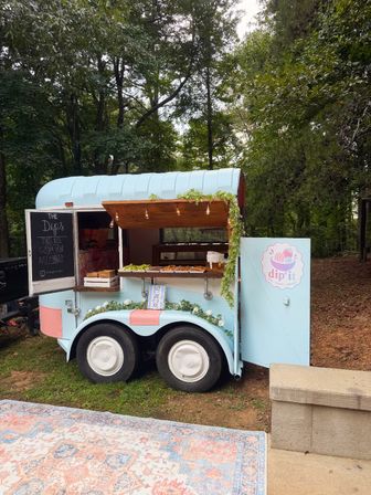 Quirky pastel-blue mobile snack trailer parked in a leafy outdoor setting, open wooden awning with string lights, chalkboard menu and trays of dips and treats on the serving counter