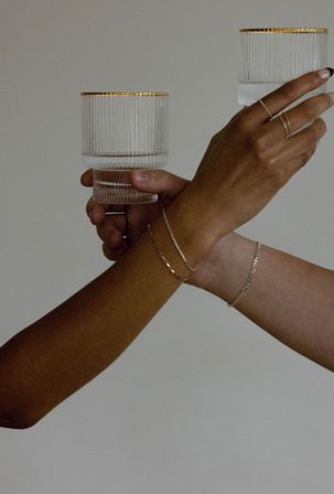 Two hands holding ribbed, gold‑rimmed glasses against a neutral backdrop, accented with delicate gold and silver bracelets and stacked rings — minimalist modern drinkware and jewelry styling.