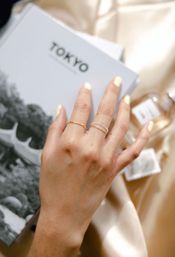 Close-up of a hand with pale yellow manicure wearing delicate gold and silver stacking rings resting on a 'TOKYO' book and satin — minimalist travel-inspired jewelry shot.