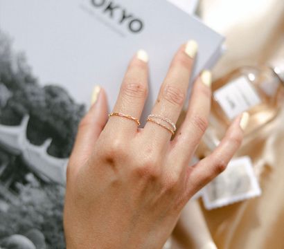 Close-up of a hand with pale yellow manicure wearing delicate gold and silver stacking rings resting on a 'TOKYO' book and satin — minimalist travel-inspired jewelry shot.
