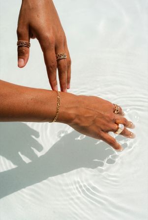 Sunlit tan hands wearing gold rings and a chain bracelet skimming clear shallow pool water, creating gentle ripples and crisp shadows.
