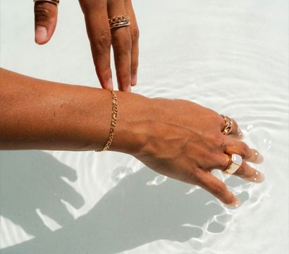 Sunlit tan hands wearing gold rings and a chain bracelet skimming clear shallow pool water, creating gentle ripples and crisp shadows.