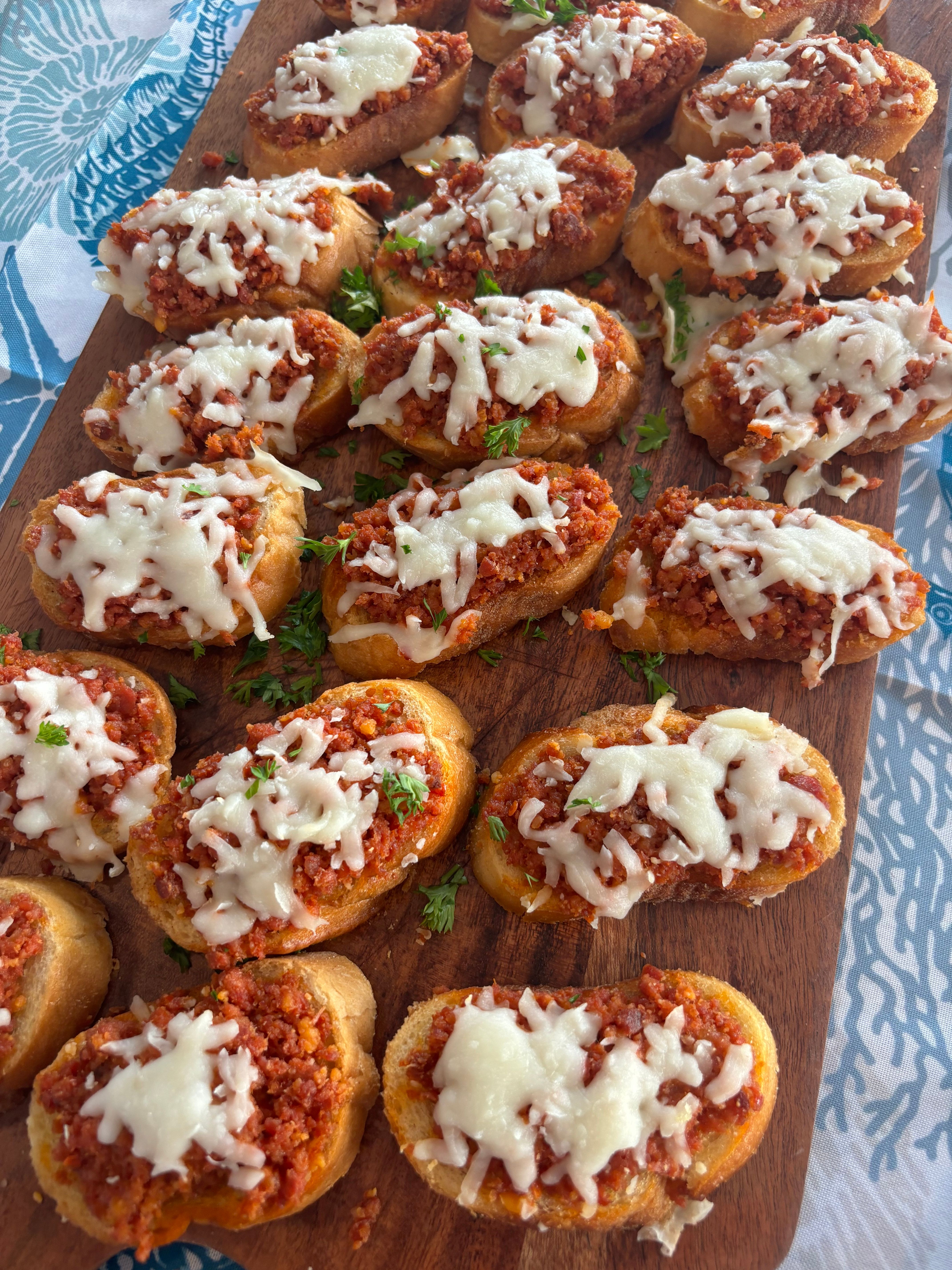 Party-ready Italian crostini: toasted baguette slices topped with savory tomato-meat ragù, melted mozzarella and parsley on a wooden serving board.