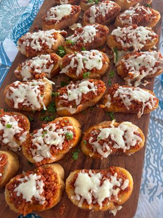 Party-ready Italian crostini: toasted baguette slices topped with savory tomato-meat ragù, melted mozzarella and parsley on a wooden serving board.