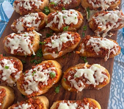 Party-ready Italian crostini: toasted baguette slices topped with savory tomato-meat ragù, melted mozzarella and parsley on a wooden serving board.