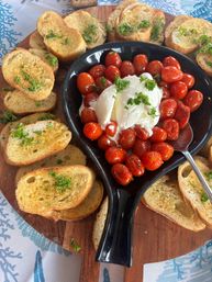 Roasted cherry tomatoes and creamy burrata in a black dish surrounded by garlic‑herb crostini on a wooden serving board — Mediterranean‑style appetizer for sharing.