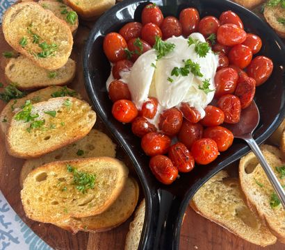Roasted cherry tomatoes and creamy burrata in a black dish surrounded by garlic‑herb crostini on a wooden serving board — Mediterranean‑style appetizer for sharing.