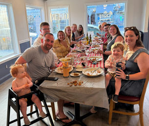 Large multigenerational group enjoying a casual family meal at a long checkered-tablecloth table in a bright local restaurant, adults and young children smiling amid plates, wine bottles and shared dishes.