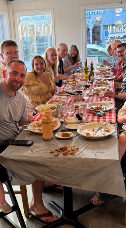 Large multigenerational group enjoying a casual family meal at a long checkered-tablecloth table in a bright local restaurant, adults and young children smiling amid plates, wine bottles and shared dishes.
