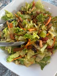 Crunchy romaine salad tossed with shredded carrots, grape tomatoes, dried cranberries and candied pecans in a creamy dressing, served in a white bowl with metal tongs