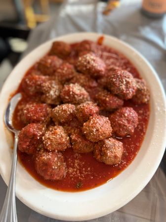 Heaping white platter of Italian meatballs in chunky marinara sauce, sprinkled with grated Parmesan and served with a spoon