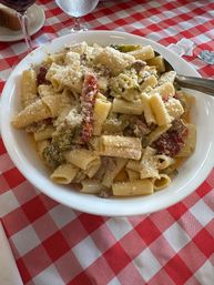 Hearty rigatoni pasta tossed in creamy sauce with grated Parmesan, sun-dried tomatoes, broccoli and sausage in a white bowl on a red-and-white checkered restaurant tablecloth with wine and water glasses visible.