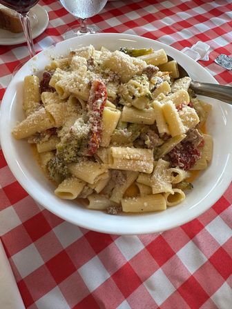 Hearty rigatoni pasta tossed in creamy sauce with grated Parmesan, sun-dried tomatoes, broccoli and sausage in a white bowl on a red-and-white checkered restaurant tablecloth with wine and water glasses visible.