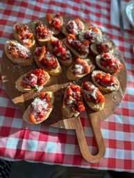Wooden board of Italian-style tomato bruschetta appetizers on toasted baguette slices topped with chopped cherry tomatoes, basil and grated cheese, on a red-and-white checkered tablecloth