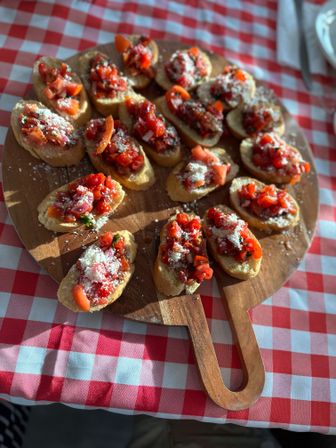 Wooden board of Italian-style tomato bruschetta appetizers on toasted baguette slices topped with chopped cherry tomatoes, basil and grated cheese, on a red-and-white checkered tablecloth