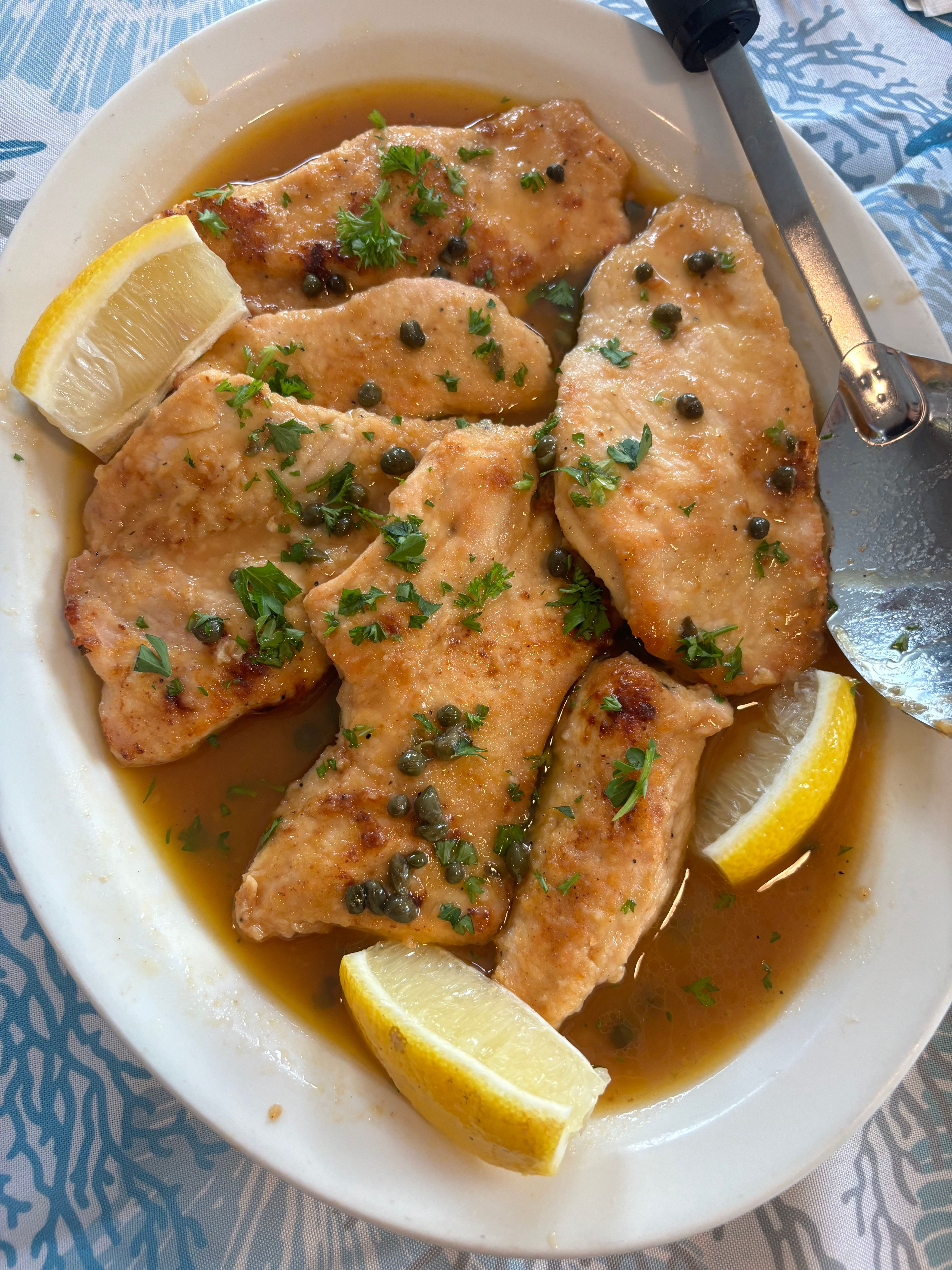 Close-up of Italian-style chicken piccata — pan-seared chicken cutlets in zesty lemon-caper butter sauce, garnished with chopped parsley and lemon wedges on a white plate.