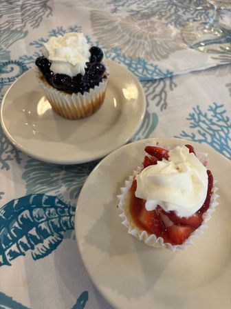 Two mini fruit cupcakes—one blueberry-topped and one strawberry-topped with whipped cream—served on white saucers over a blue coral beach-themed tablecloth.