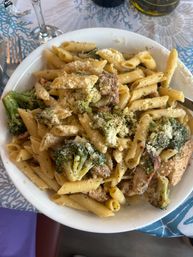 Hearty bowl of penne pasta with broccoli and grilled chicken in a garlic-herb sauce, topped with grated Parmesan, served in a white bowl on a blue patterned table.