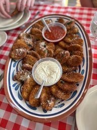 Large ceramic platter of crispy breaded fried zucchini rounds sprinkled with Parmesan and herbs, served with marinara and creamy garlic dipping sauces on a red-and-white checkered tablecloth.