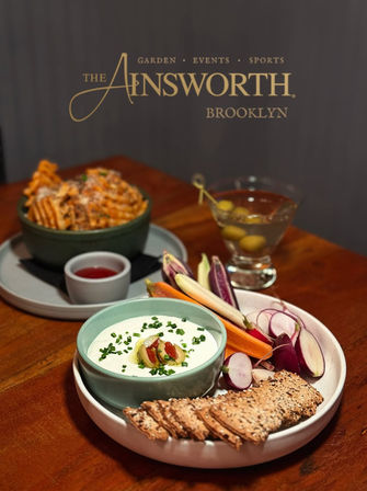 Appetizer platter on a wooden table in an upscale restaurant: creamy herbed dip topped with olives and chives, seeded crackers, radishes and baby carrots, waffle fries with dipping sauce in the background and a martini with olives.