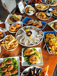 Colorful seafood spread on a wooden outdoor table: oysters on ice with cocktail sauce and lemon, fried calamari, crab cakes, mussels, fries and onion rings — seaside restaurant appetizers.