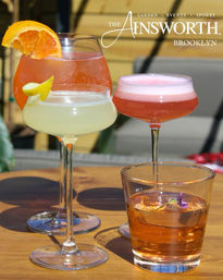 Four colorful cocktails on a sunny outdoor patio table in Brooklyn — orange spritz with an orange slice, pale citrus coupe with a lemon twist, pink frothy cocktail, and an amber lowball garnished with an edible flower.