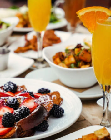 Brunch table with French toast dusted in powdered sugar and topped with strawberries and blackberries, a bowl of seasoned breakfast potatoes, and orange mimosas garnished with orange slices.