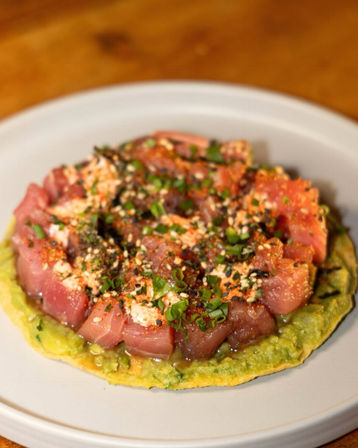Ahi tuna poke tostada — diced raw tuna seasoned with sesame, chives and spices on smashed avocado over a crispy tortilla, served on a white plate.