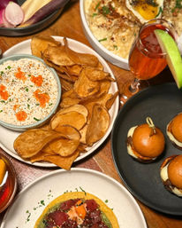 Gourmet appetizer spread on a wooden table — hand-cut potato chips with a creamy roe-topped dip, mini brioche sliders, a tuna poke tostada with avocado, and a sparkling apple-garnished cocktail.