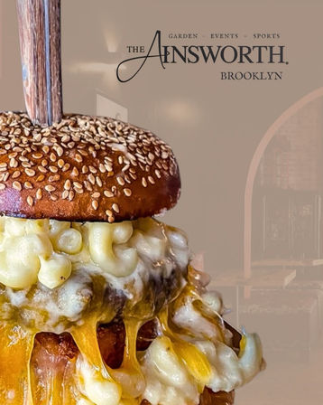 Close-up of a decadent sesame-seed burger stacked with creamy macaroni-and-cheese and gooey melted cheddar dripping down, secured with a wooden skewer against a soft neutral background.