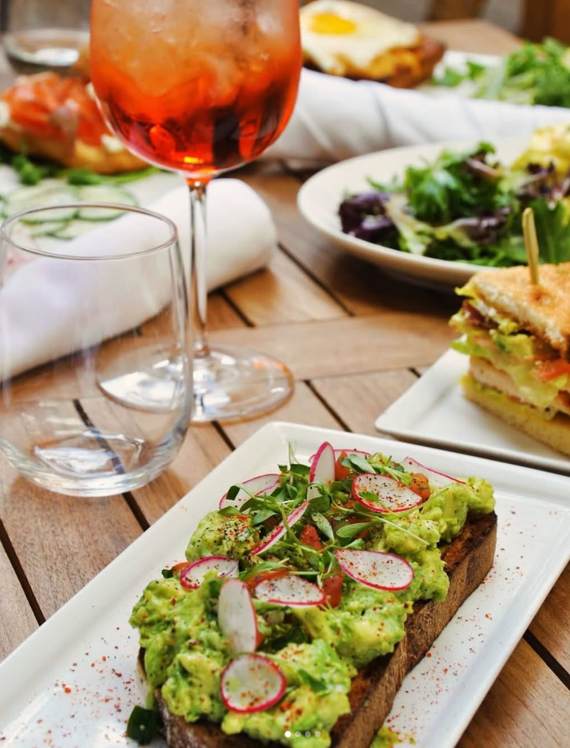 Brunch spread: avocado toast with sliced radishes and microgreens on toasted sourdough, iced orange cocktail, mixed green salad and sandwich on a wooden patio table.