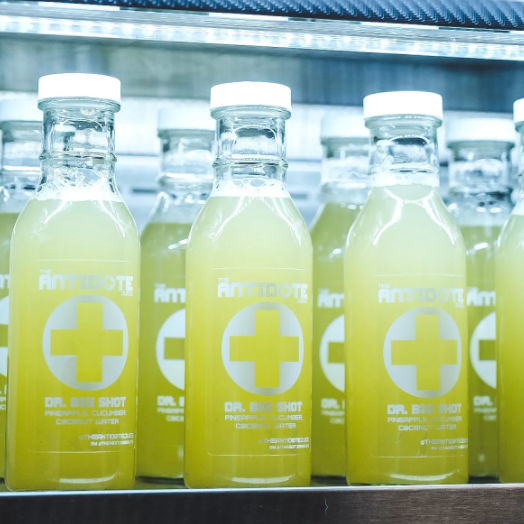 Bright green cold-pressed juice shot bottles in clear glass with white caps and cross logo on a refrigerated shelf