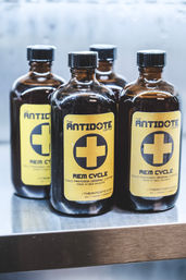 Four amber glass bottles of cold-pressed coffee with yellow labels featuring a bold medical-style cross, lined up on a stainless-steel shelf.