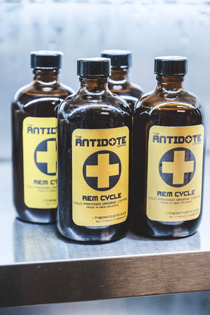 Four amber glass bottles of cold-pressed coffee with yellow labels featuring a bold medical-style cross, lined up on a stainless-steel shelf.