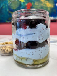 Close-up of a layered yogurt parfait in a glass jar with banana base, chia-seed yogurt swirls, fresh blueberries and a small cup of granola on a white table against a colorful backdrop.