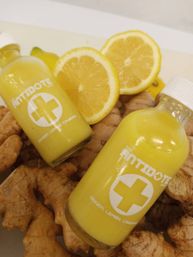 Two small yellow ginger-lemon wellness shot bottles with white caps nestled on fresh ginger root and lemon halves — bright immune-boosting juice.