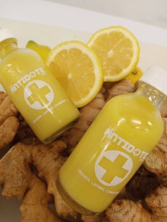 Two small yellow ginger-lemon wellness shot bottles with white caps nestled on fresh ginger root and lemon halves — bright immune-boosting juice.