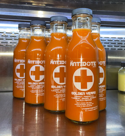 Bright orange cold-pressed turmeric-carrot juice in glass bottles with white cross logo and ingredient list (carrot, pineapple, turmeric, ginger, ginseng, black pepper) lined up on a stainless-steel refrigerated shelf