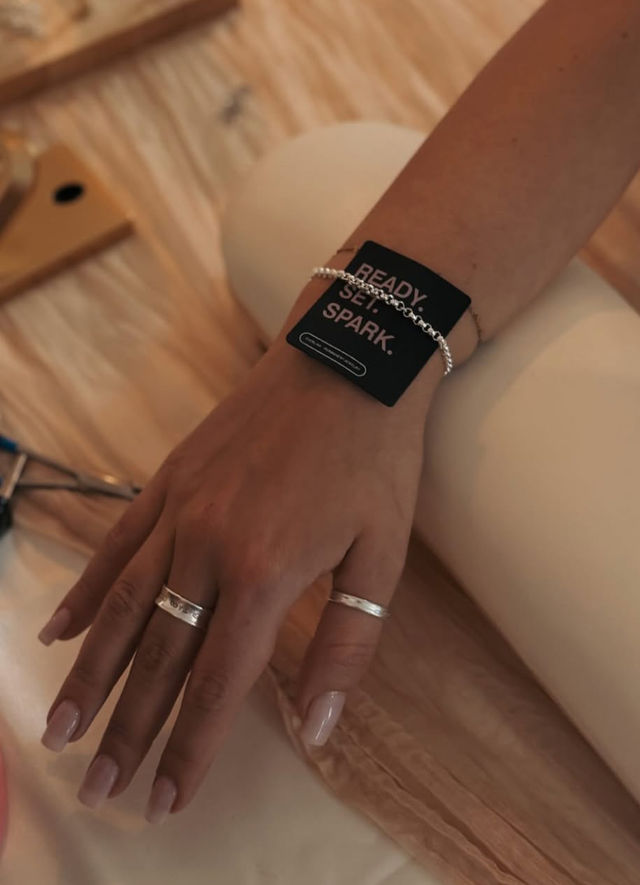 Close-up of a manicured hand with nude nails wearing silver rings and a chain bracelet with a black tag reading READY. SET. SPARK. on a warm beige surface