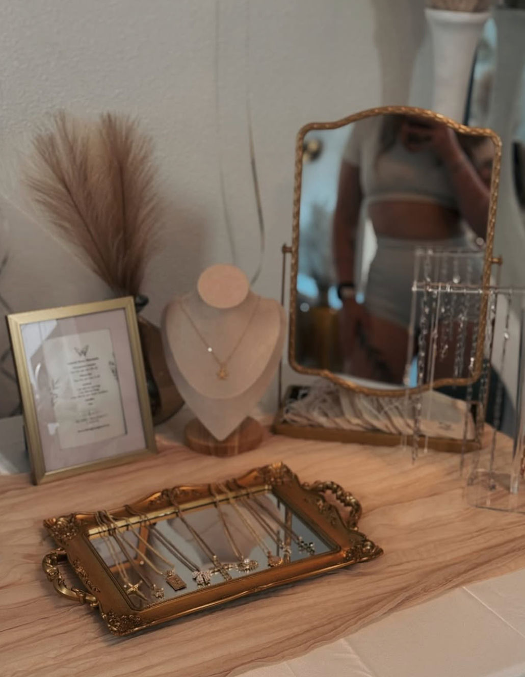 Boho vanity scene on a wooden dresser: ornate gold tray displaying delicate necklaces, a necklace bust with star pendant, a gold-framed standing mirror with a blurred reflection, dried pampas grass and a framed print.