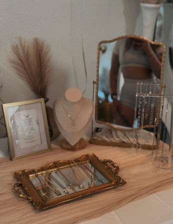 Boho vanity scene on a wooden dresser: ornate gold tray displaying delicate necklaces, a necklace bust with star pendant, a gold-framed standing mirror with a blurred reflection, dried pampas grass and a framed print.