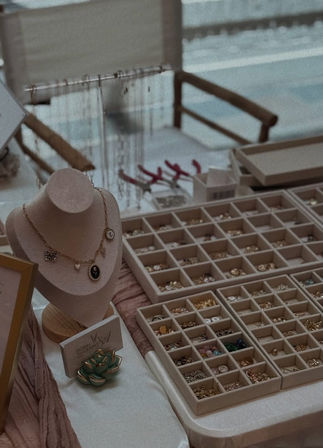 Velvet necklace bust displaying a charm necklace beside organized trays of rings and earrings on a market table, with jewelry tools and a small ceramic succulent.